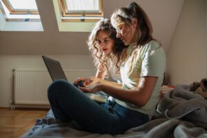 Two sisters sit in their room, focused on a laptop. They collaborate on a project, typing and discussing ideas as they work together in a relaxed environment.